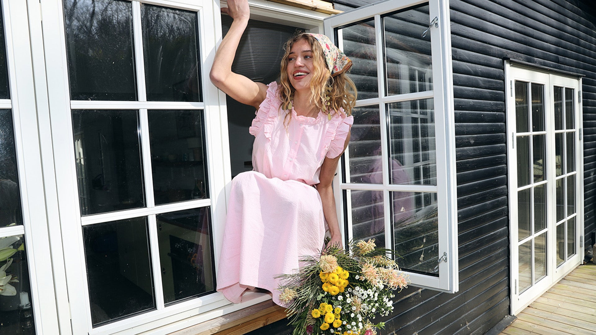 Kvinde i en lys rosa maxikjole siddende i et åbent vindue ved et sort sommerhus med en buket vilde blomster i hånden.