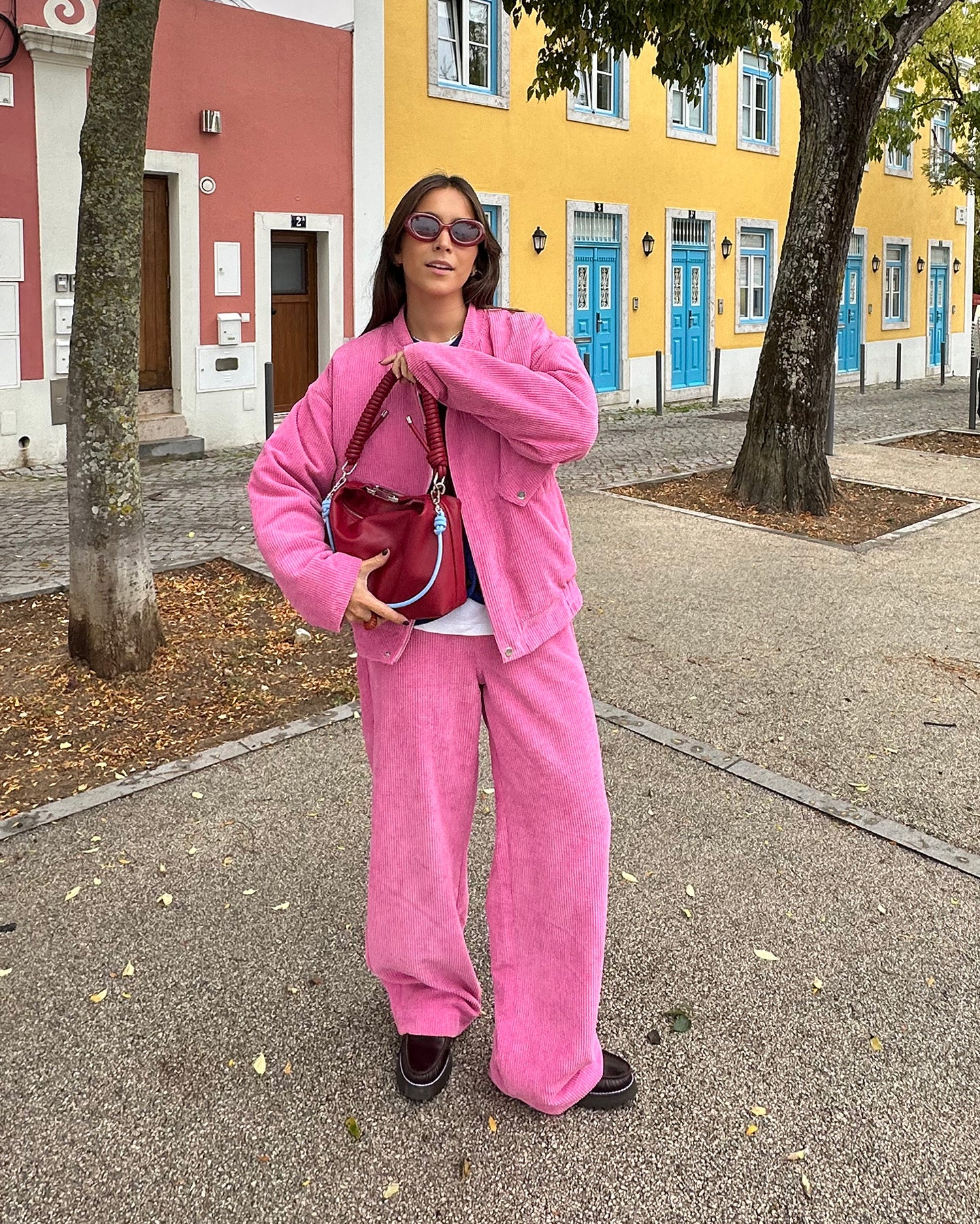 En kvinde står udendørs iført et pink corduroy sæt med jakke og brede bukser. Hun bærer en rød taske, har mørke solbriller på og står foran farverige huse med blå døre.