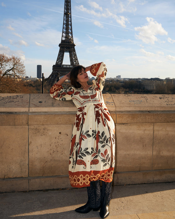 Kvinde i en luftig midi kjole med et detaljeret print i røde og creme farver, stående foran Eiffeltårnet i Paris. Kjolen har lange ærmer, en afslappet pasform og dekorative kanter, stylet med sorte støvler for et moderne og ubesværet look.