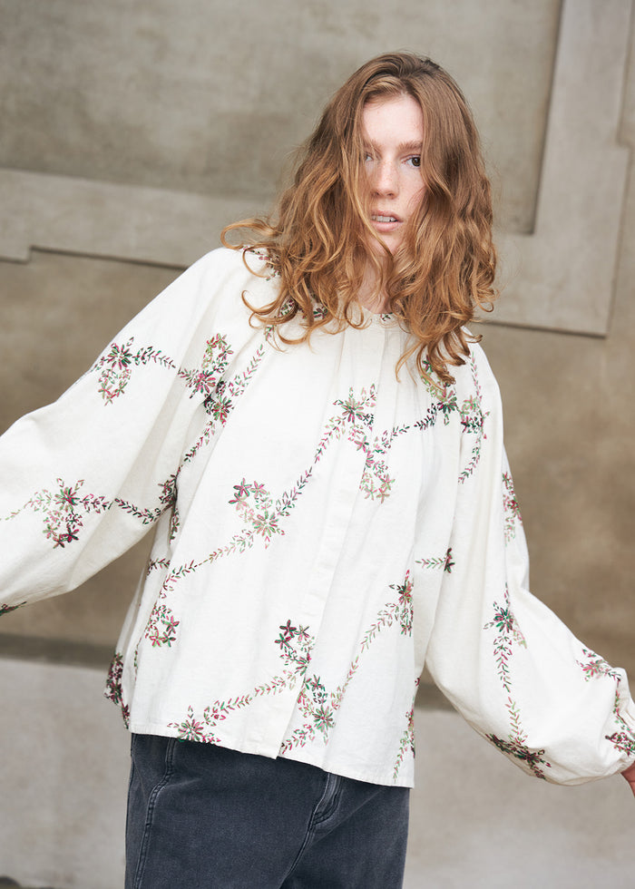 Kvinde iført hvid bluse med farverige blomsterbroderier og voluminøse ærmer, stylet med mørke denimjeans. En feminin hverdagsstyle med dekorative detaljer.