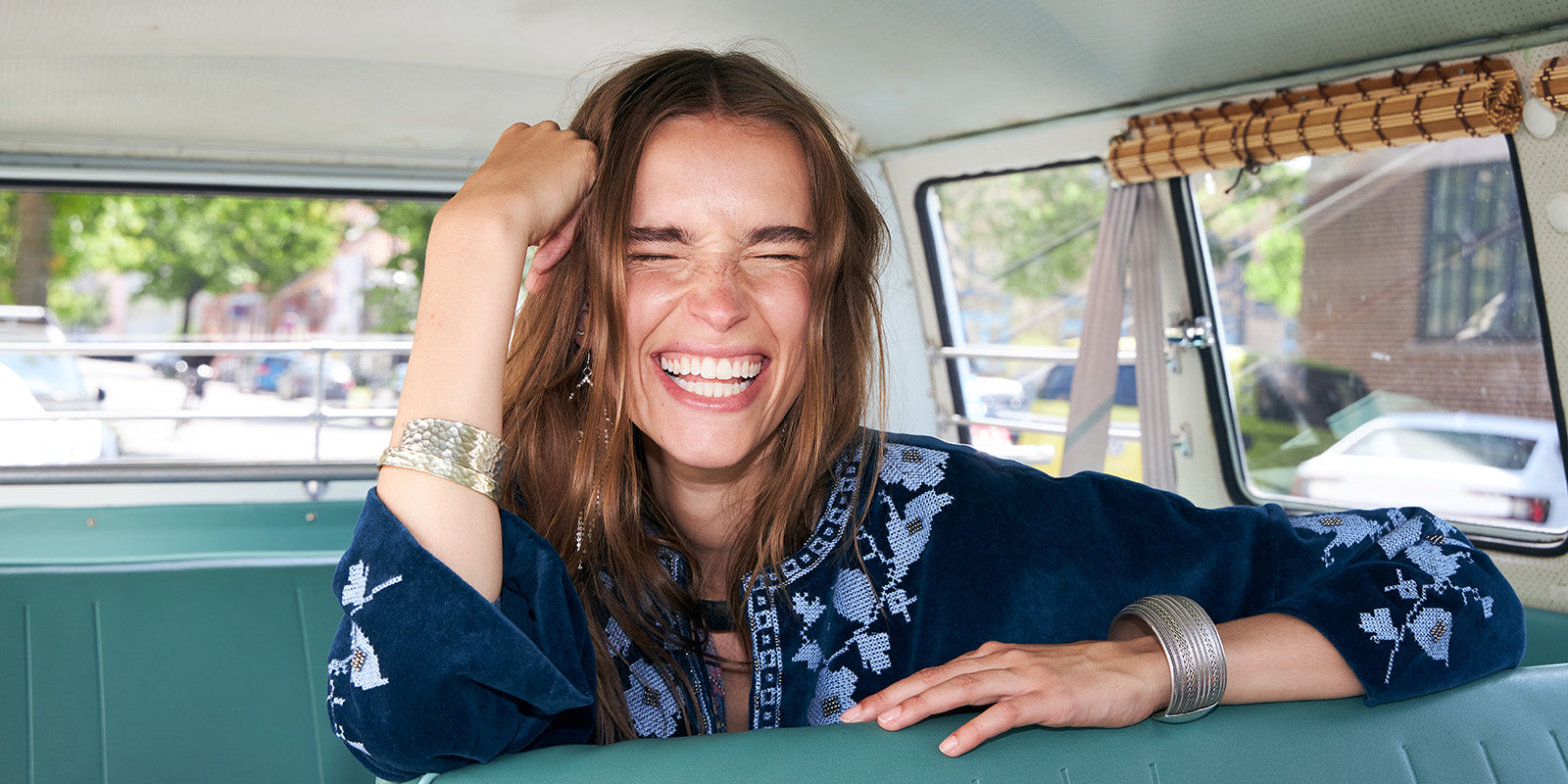 Smilende kvinde med langt brunt hår sidder i en vintage minibus. Hun læner sig afslappet over sædet og griner med lukkede øjne. Hun bærer en mørkeblå jakke med lyseblå broderede detaljer og sølvfarvede armbånd. Sollyset strømmer ind gennem vinduerne og skaber en varm, sommerlig stemning.