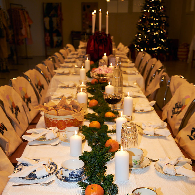 Langt julepyntet bord med tændte stearinlys, grangrene, appelsiner og mønstret porcelæn. I baggrunden lyser et juletræ.