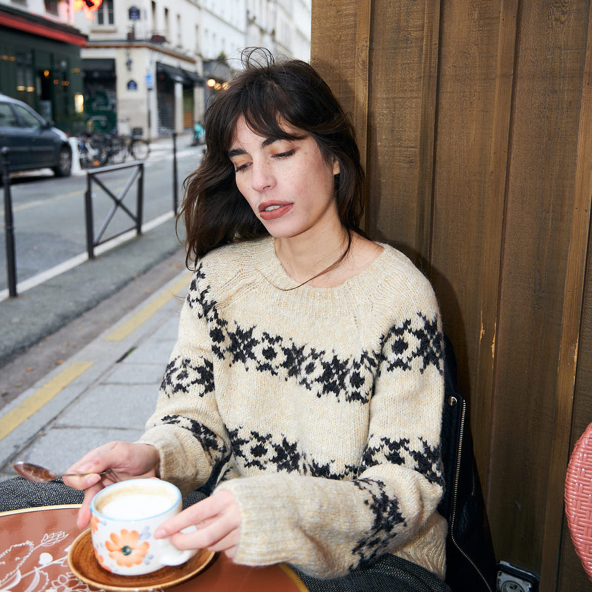 Model iført cremefarvet Lollys Laundry striktrøje med mørkt nordisk mønster, siddende udendørs på en café med en kop kaffe.