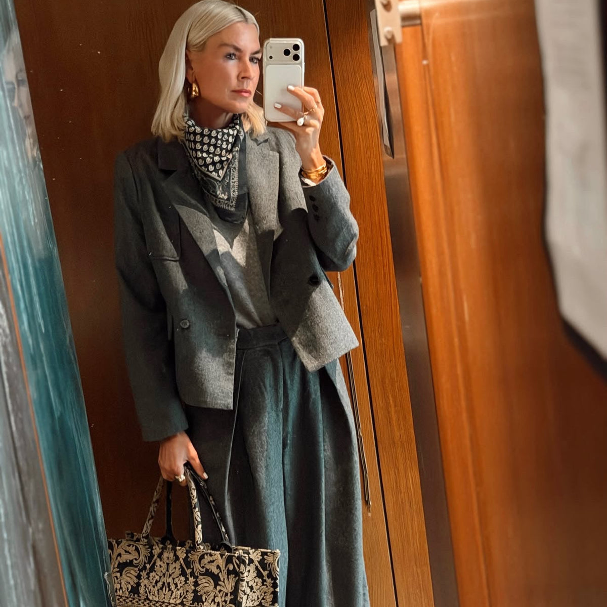 En kvinde tager en spejl-selfie indendørs foran vægge med træpaneler hun er iført en grå blazer, et mønstret tørklæde og grå bukser med brede ben, og hun holder en sort- og beige-mønstret totebag.