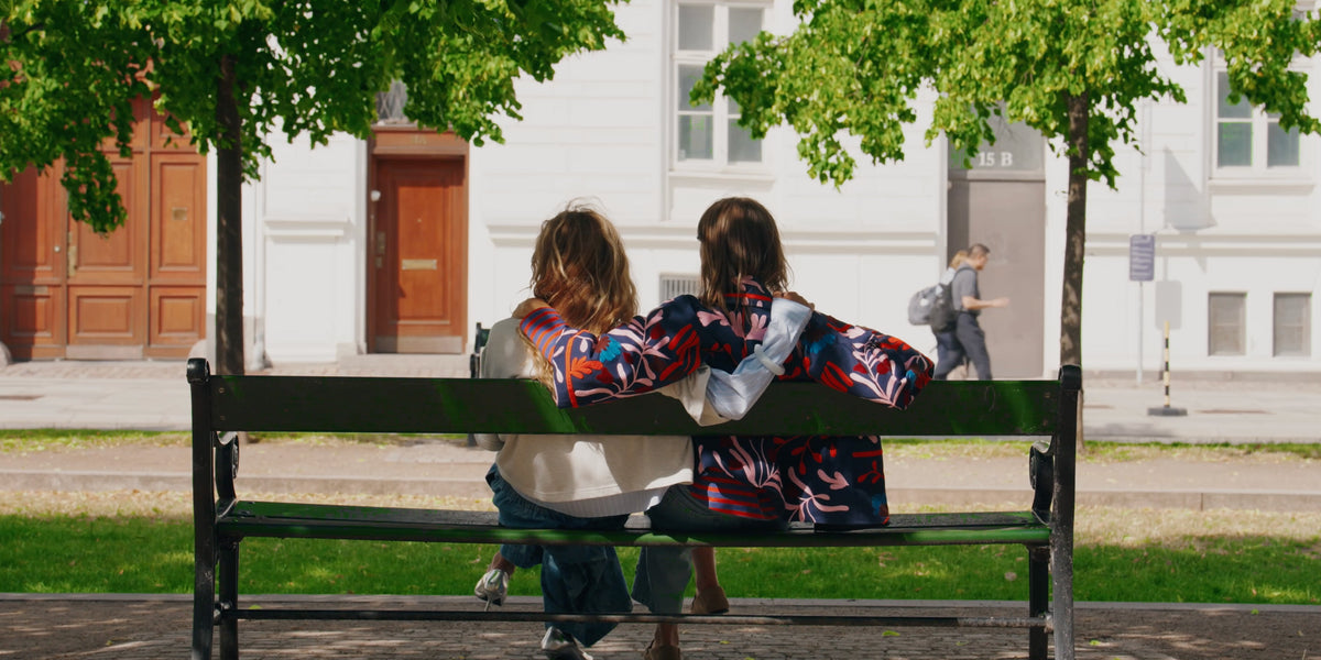 To kvinder sidder med armene om hinanden på en bænk i København, iført farverige styles fra Lollys Laundry.