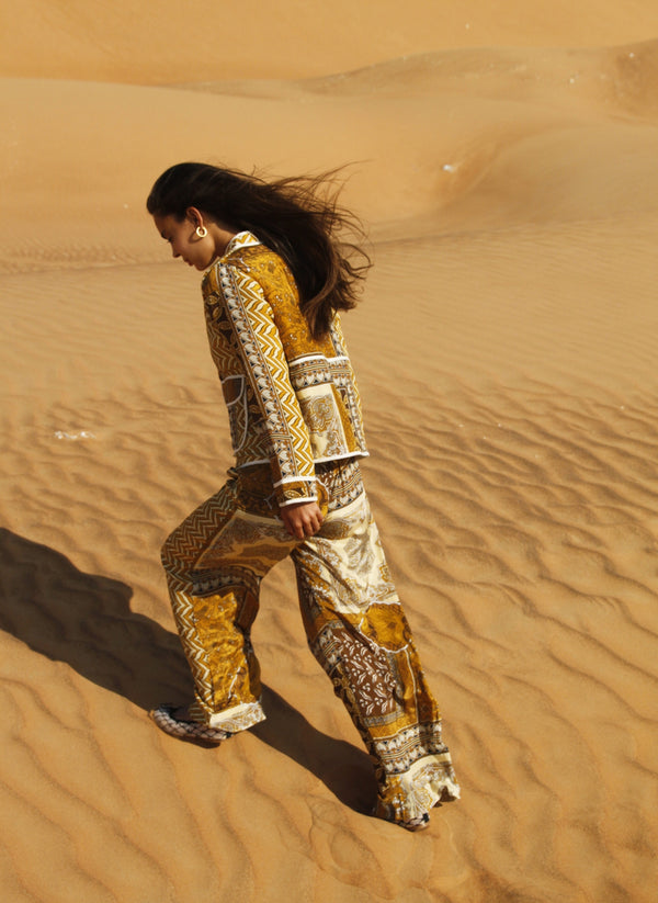 Kvinde går op ad en sandklit i ørkenen iført et matchende sæt med jakke og bukser i varme gyldne og brune nuancer med patchworkmønster. Hendes hår blæser i vinden, og lyset fremhæver teksturerne i sandet og tøjets detaljer.
