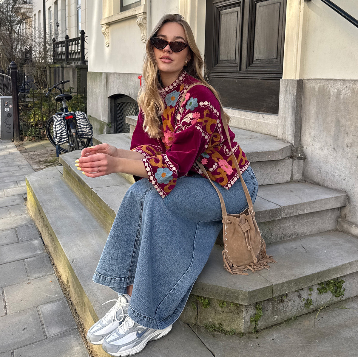 Kvinde iført bordeaux jakke fra Lollys Laundry med farverige blomsterbroderier og dekorative kanter, stylet med vide jeans, sneakers og brun skuldertaske.