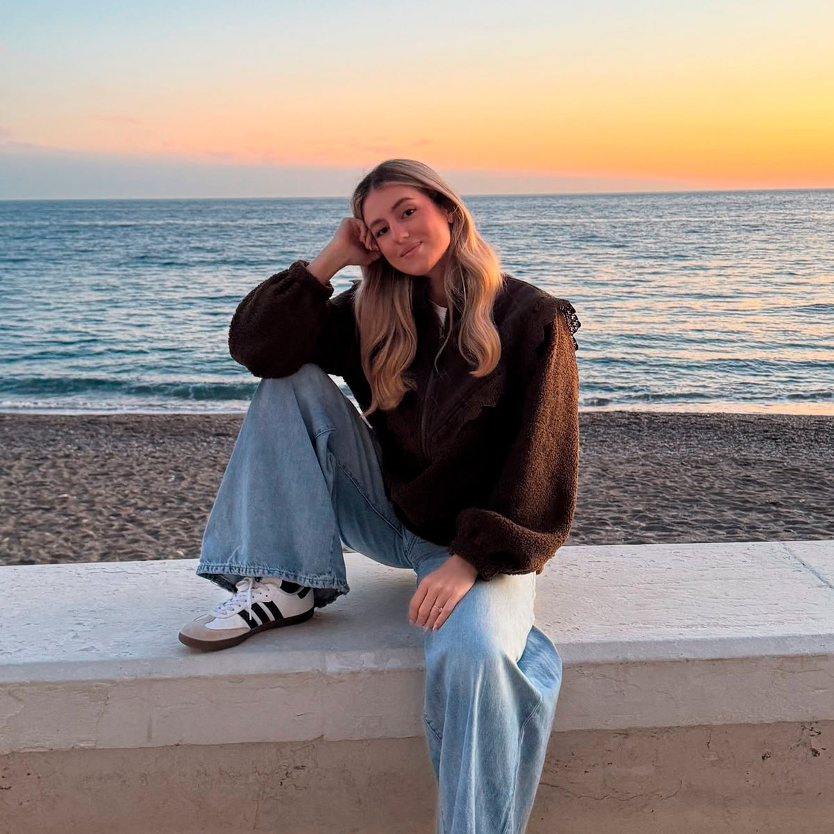 Kvinde iført mørkebrun teddyjakke med blød struktur, lyseblå jeans og sneakers, siddende ved stranden i solnedgangen.
