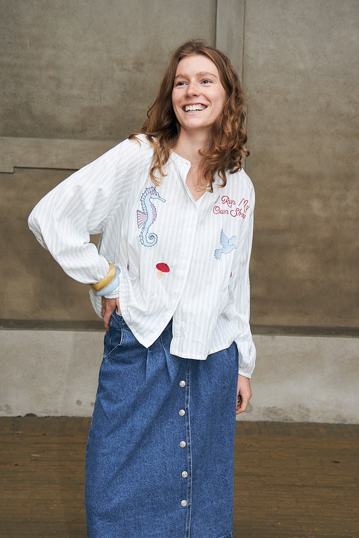 Kvinde iført hvid broderet skjorte med farverige motiver og lang denimnederdel. Et afslappet og feminint hverdagslook, perfekt til sæsonens garderobe.