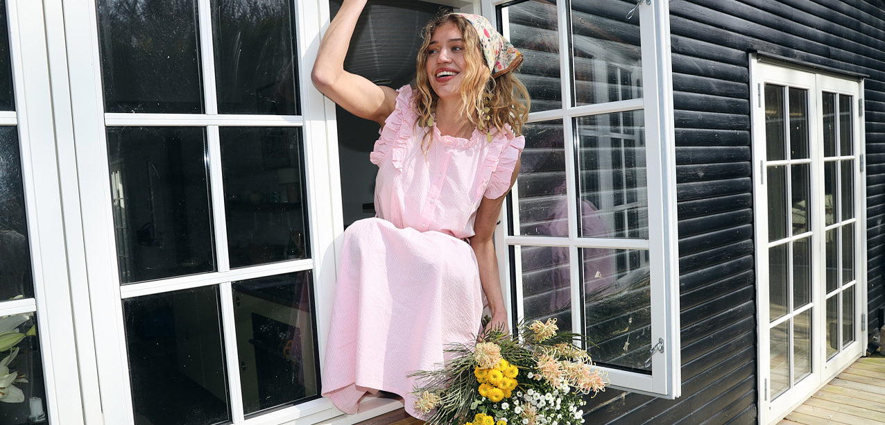 Kvinde i en lyserød kjole med flæsedetaljer og blomstrede tørklæde siddende i et åbent vindue med en blomsterbuket nedenfor.