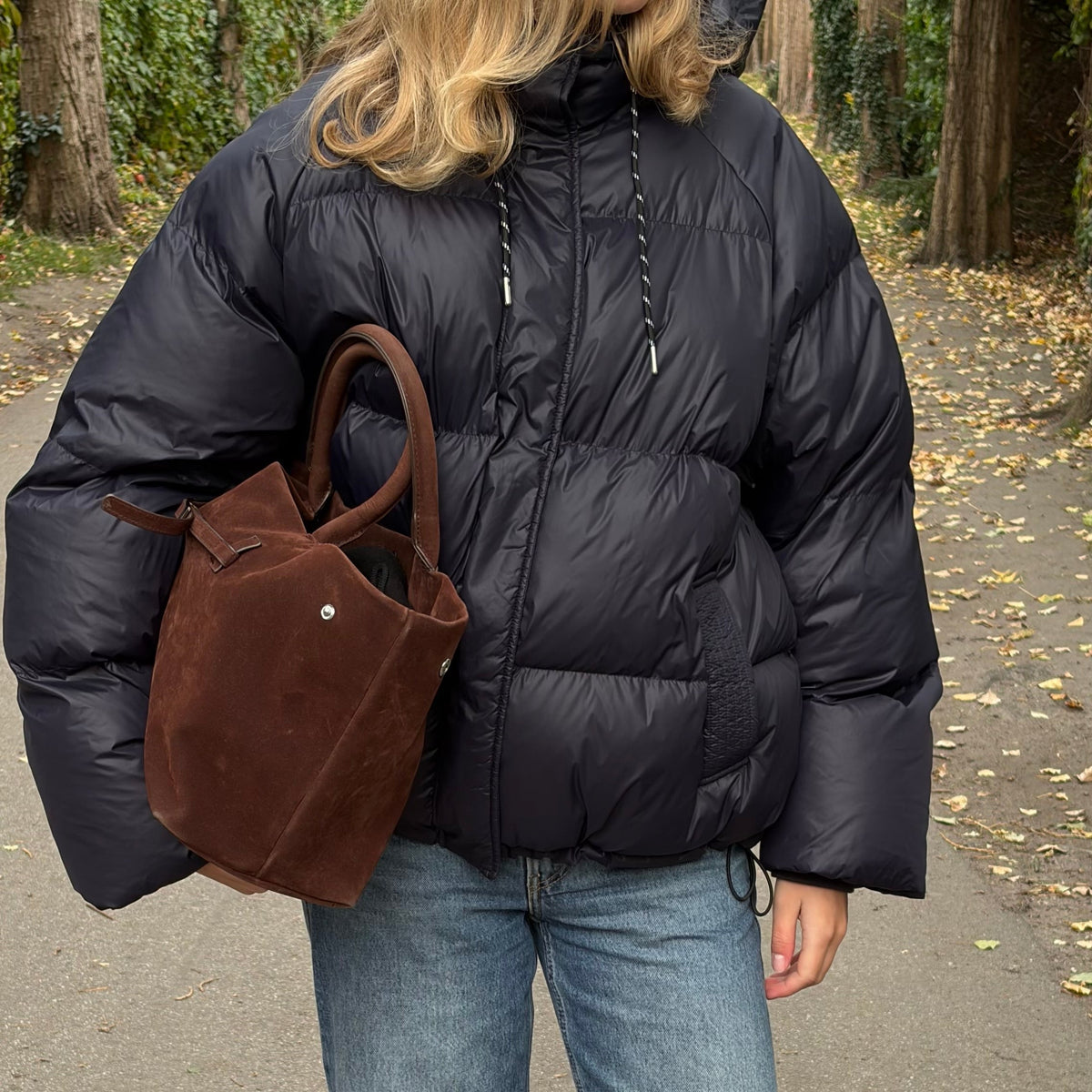 En kvinde står udendørs på en flisebelagt sti iført sort dunjakke, blå jeans og brune sko, mens hun bærer en brun ruskindstaske.