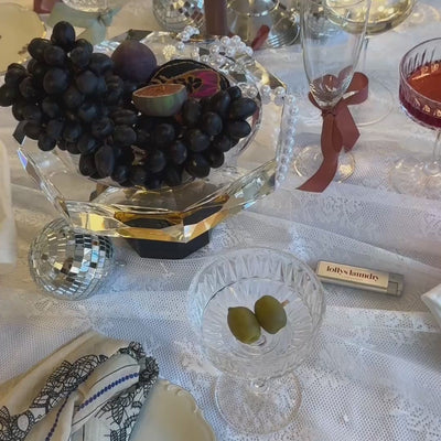 Festligt dækket julebord med vindruer, frugt, krystalglas, diskokugler og fine dekorative detaljer på en hvid dug.