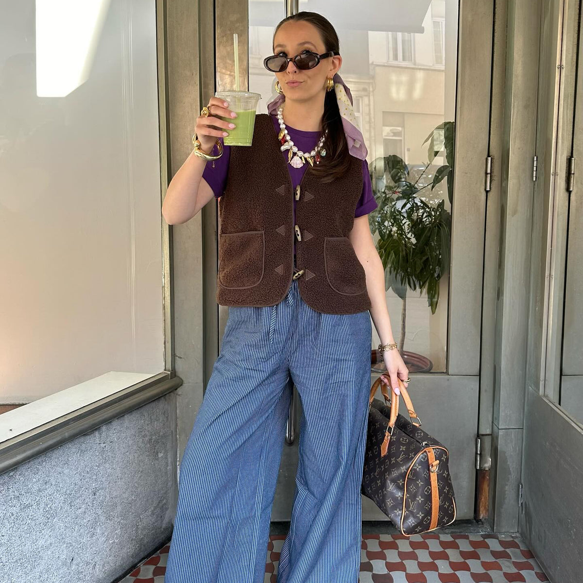 Kvinde iført brun vest over lilla T-shirt og blåstribede bukser med vide ben, stylet med solbriller, smykker og mønstret taske.